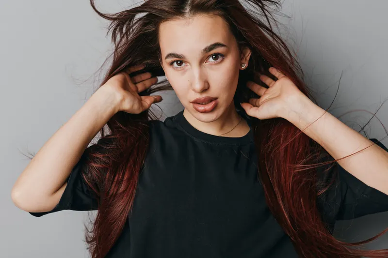 Young woman with long hair and natural makeup posing with hands near head against neutral background. Casual style, confident expression and indoor portrait with soft lighting.