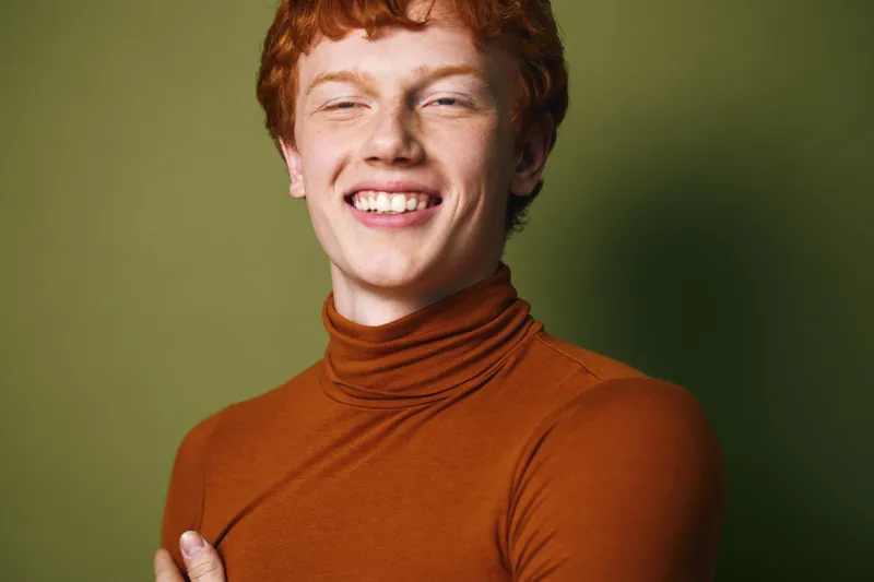 Young man with red hair and freckles wearing brown turtleneck smiling with eyes closed in front of green background. Portrait of relaxed confident male model with casual style indoors.