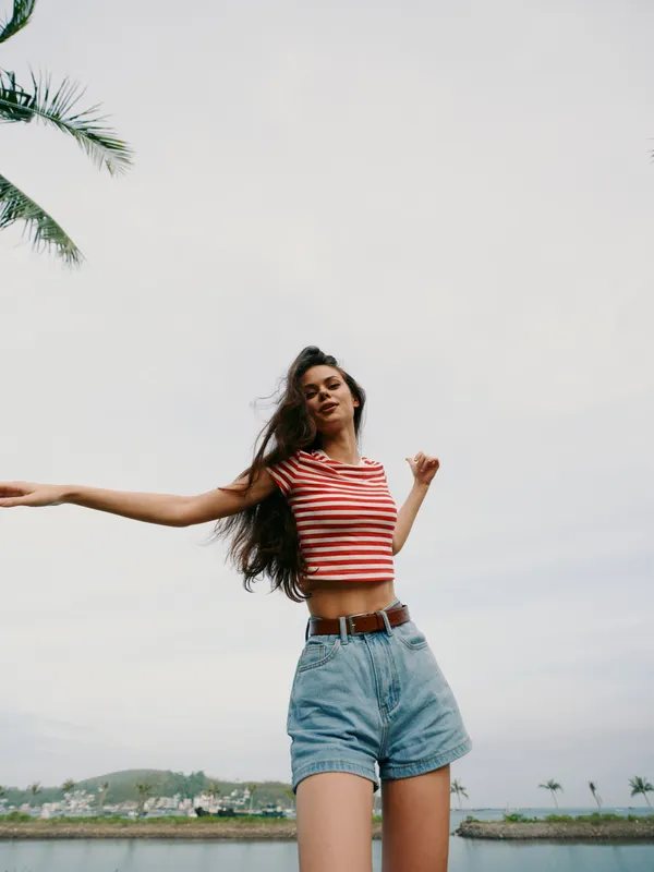 A young woman stands outdoors in a striped crop top and denim shorts, with a palm tree and cloudy sky creating a relaxed summer mood.