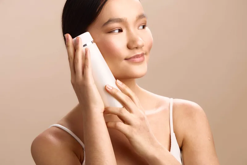 Asian woman holds a bottle to her ear in a soft studio setting, suggesting skincare use. She smiles lightly, creating a calm, confident mood and a modern beauty aesthetic.