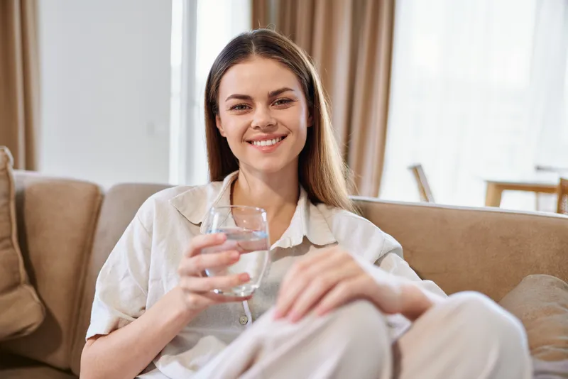 Woman smile glass water sofa home relaxed portrait of a young woman sitting on a couch holding a glass of water, casual indoor lifestyle and hydration in bright living room