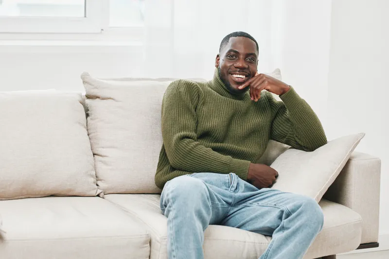 A smiling African man in a green turtleneck sweater seated on a light couch in a bright, modern living room, exuding a cozy and relaxed atmosphere