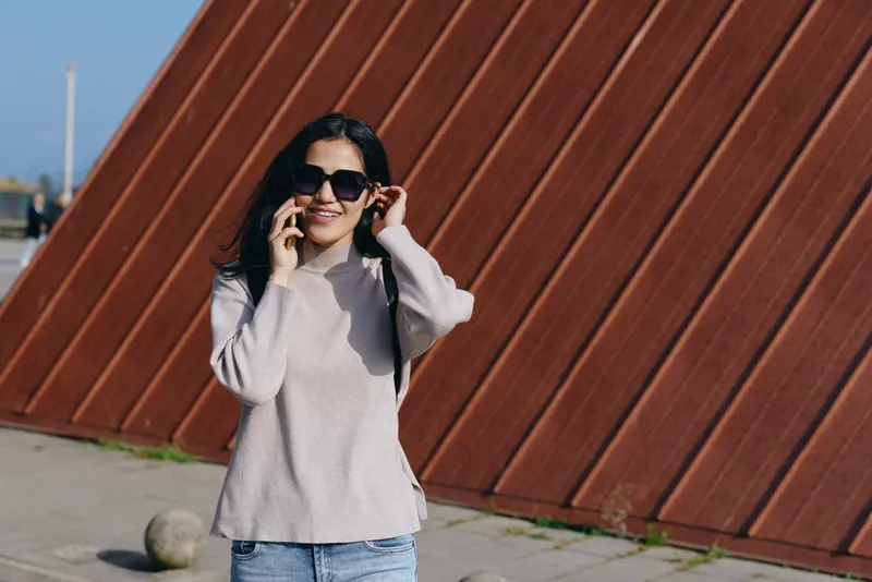 Woman in a light sweater and jeans chats on her phone outdoors, wearing sunglasses beside a bold rust wall. Casual urban style, backpack straps, sunny day, candid street moment.