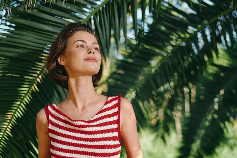 A cheerful woman stands outdoors among lush palm leaves, wearing a striped top. She gazes into the distance with a confident, relaxed smile, creating a bright, natural lifestyle moment.