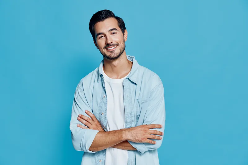 Smiling young man with crossed arms looking at the camera, wearing light blue shirt over white t-shirt, isolated on bright blue background. People lifestyle concept