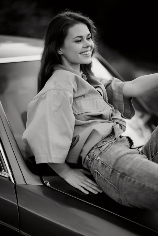 Happy young woman in oversized shirt sitting on vintage car, capturing joy and carefree spirit in a black and white portrait, ideal for lifestyle and fashion themes.
