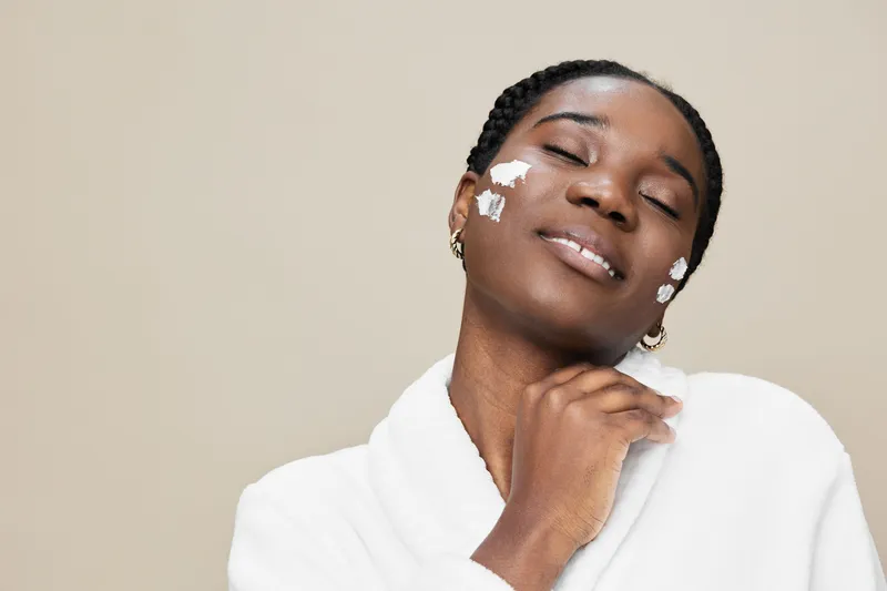 A young Black woman enjoying a skincare routine with cream on her face, wearing a cozy white robe, smiling with confidence against a neutral backdrop