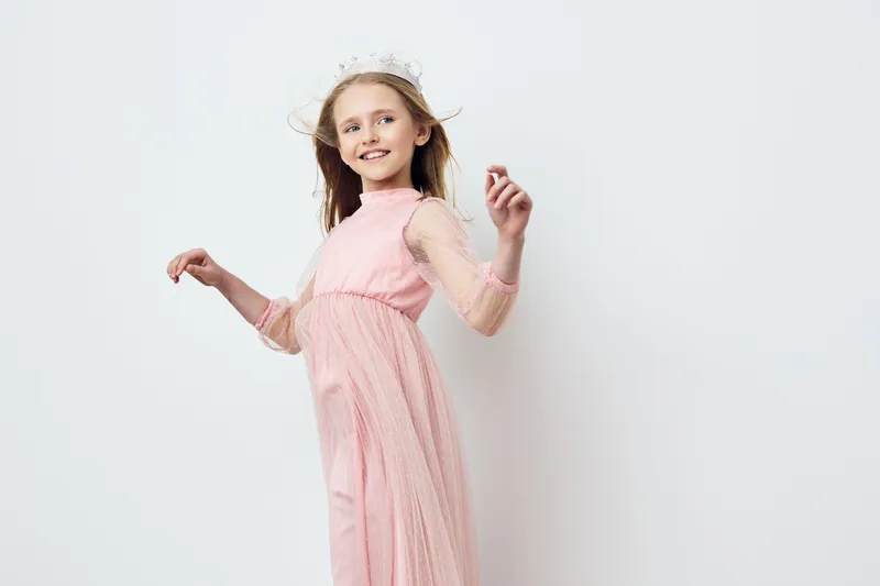 A joyful girl in a flowing pink dress poses elegantly against a minimalist white background, exuding happiness and innocence with a charming flower crown