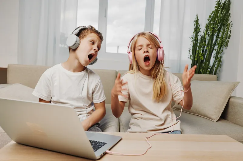Two children wearing headphones sing passionately in front of a laptop at home, expressing joy and creativity in a cozy living room with natural light and plants.