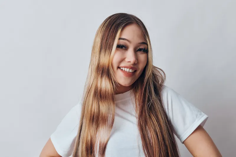 Portrait of confident woman with long hair smiling and standing with hands on hips against grey background. Young female in casual white t shirt showing positive emotion and approachable attitude.
