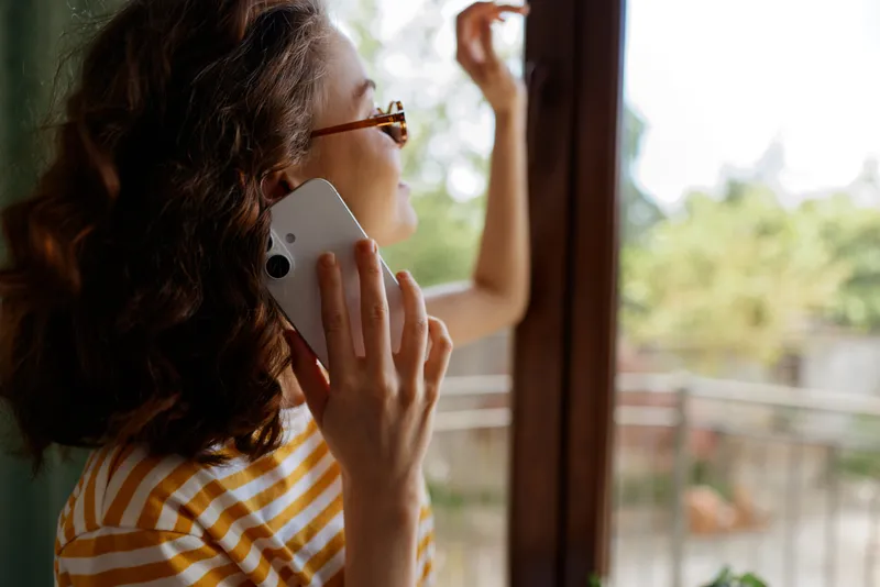 One woman with curly brown hair wearing a striped shirt talks on a mobile phone by an open window, capturing a calm, everyday home lifestyle moment and a sense of connection.