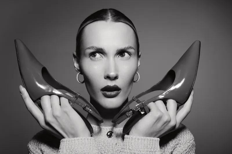 White woman with pale skin holds red high heels near face, looking sideways with serious expression. Black and white portrait in studio lighting.