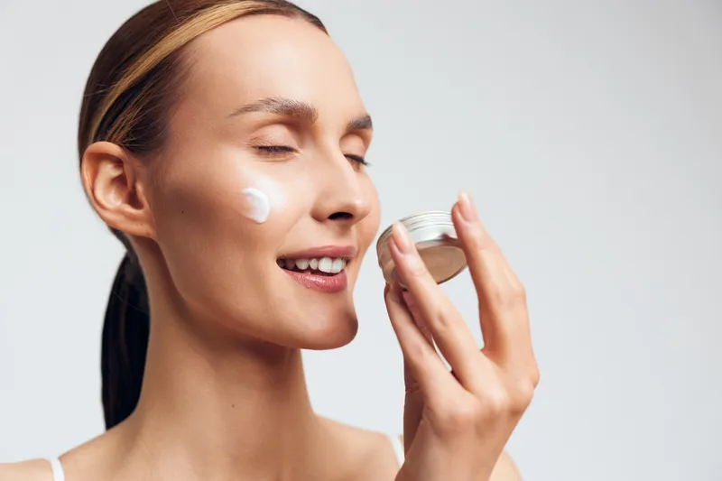 Young woman with brunette hair applies moisturizing cream on her cheek while holding a small jar in a bright studio, expressing calmness and self-care.