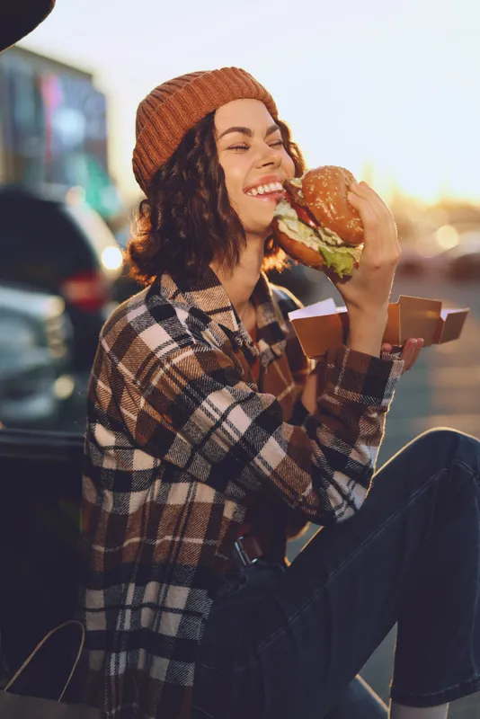 Woman eating burger with smile in candid outdoor moment, plaid jacket and beanie, authenticity and mindful living vibe, golden glow backlight and emotional storytelling lifestyle.