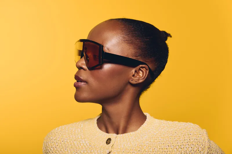 Profile of a confident black woman with dark skin wearing large sunglasses and a textured yellow sweater against a bright yellow background. Concept of fashion and style.