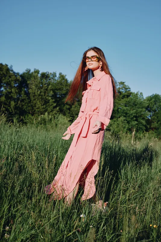 Young model in a fashionable pink dress standing in a green field against a clear blue sky, exuding confidence and joy, perfect for fashion and seasonal themes