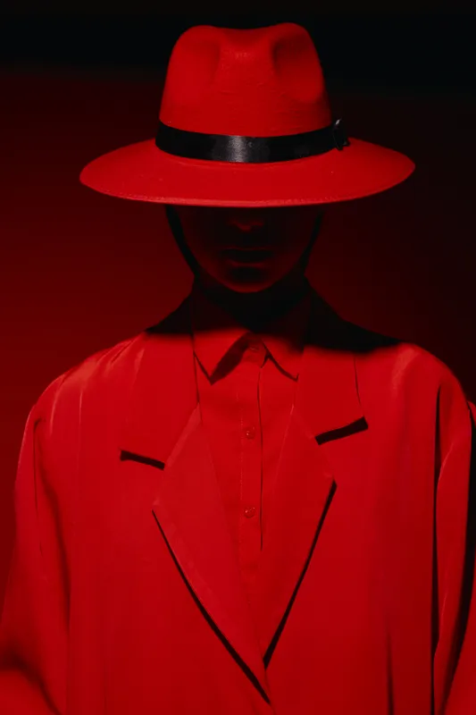 Fashion model in bold red hat and matching coat stands against dark red background with low dramatic lighting. Concept of beauty, style, and creative fashion.