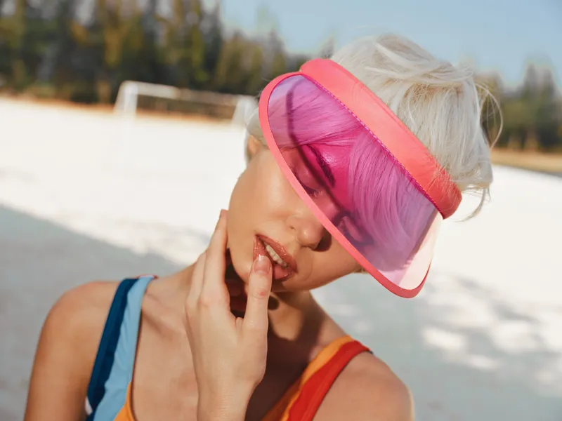 Woman wearing a bright pink visor tilts her head, gently touching her lips as sunlight washes over a sunny outdoor setting, creating a bold fashion moment and relaxed summer vibe.