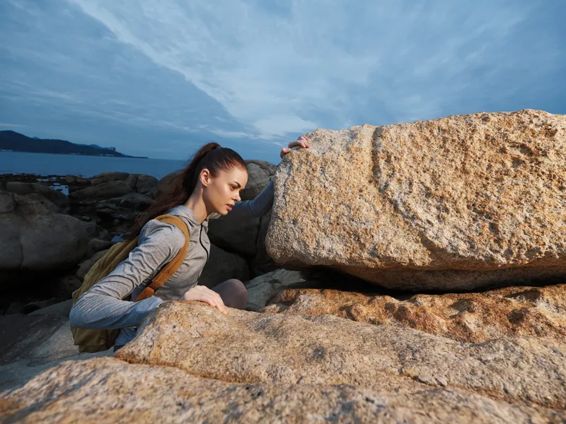 A determined young woman in activewear climbs rocky terrain by the ocean, showcasing strength and perseverance against a moody sky