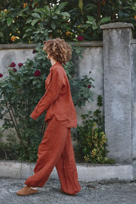 A woman in a rust colored suit walks along a garden wall, capturing a candid lifestyle moment outdoors. Warm, natural light enhances texture, color, and relaxed confidence