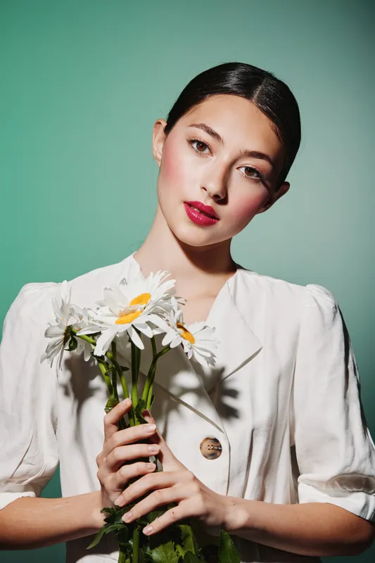 Young woman with red lipstick holds daisies looking directly into the camera. She wears a white puff-sleeve blouse. Concept for beauty creative fashion editorial style.