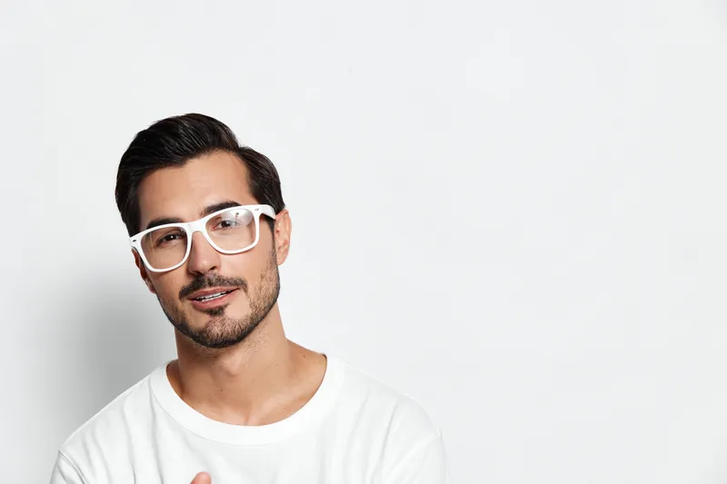 Smiling man with stylish glasses poses confidently against plain white background. Attire features casual white shirt emphasizing modern lifestyle and personality.