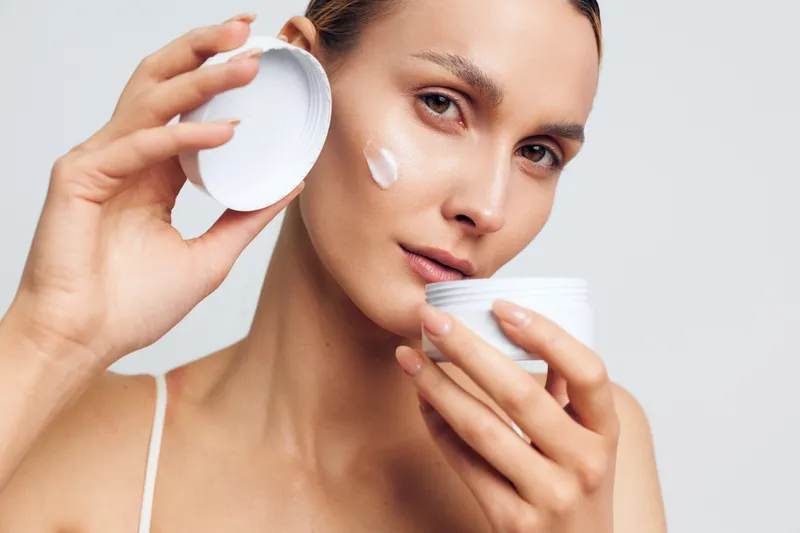 A young woman applies cream on her cheek using a white jar with a lid. The close-up portrait shows natural skin and a neutral background. Concept for skincare and beauty.