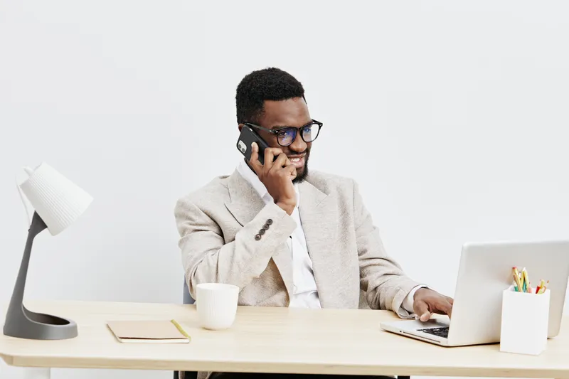 Professional young African American man working at desk using laptop and talking on mobile phone in modern office environment with minimalist decor and white background, business concept