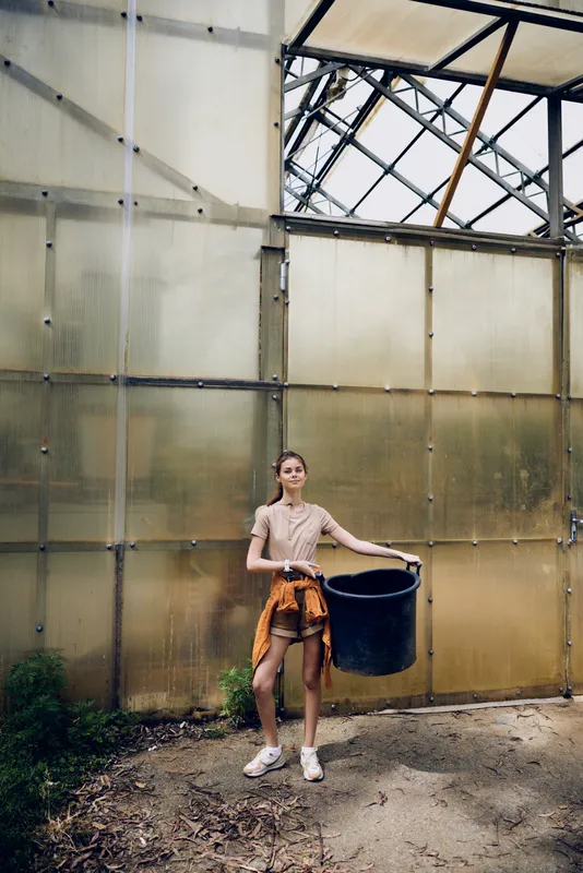 woman greenhouse bucket gardening casual plants portrait of a young female standing in a glass structure holding a black container on dirt floor, tied jacket and sneakers for outdoor hobby