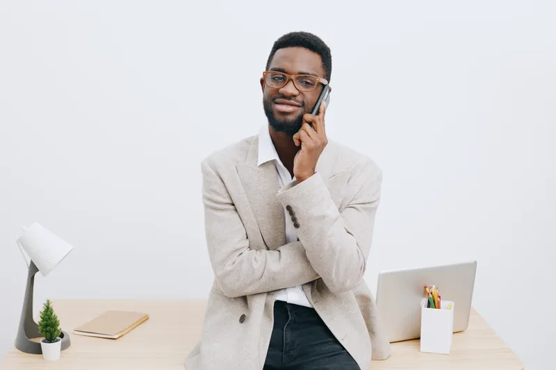 Professional man talking on mobile phone while standing at office desk, wearing glasses and beige blazer, looking confident and smiling, modern workspace with laptop, stationery and plant, indoor scen