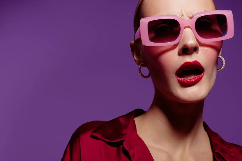 Stylish woman with pink sunglasses and red lipstick poses against purple background. Fashion concept with bold makeup, accessories, and confident expression in studio lighting.