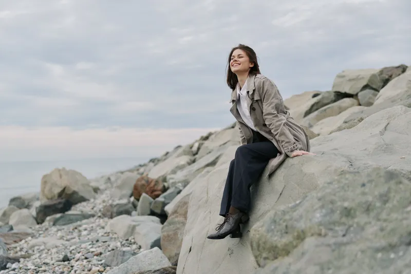 Young woman in casual clothes sits on large rocks by the sea shore, eyes closed and smiling. Calm cloudy sky in the background creates peaceful outdoor mood.