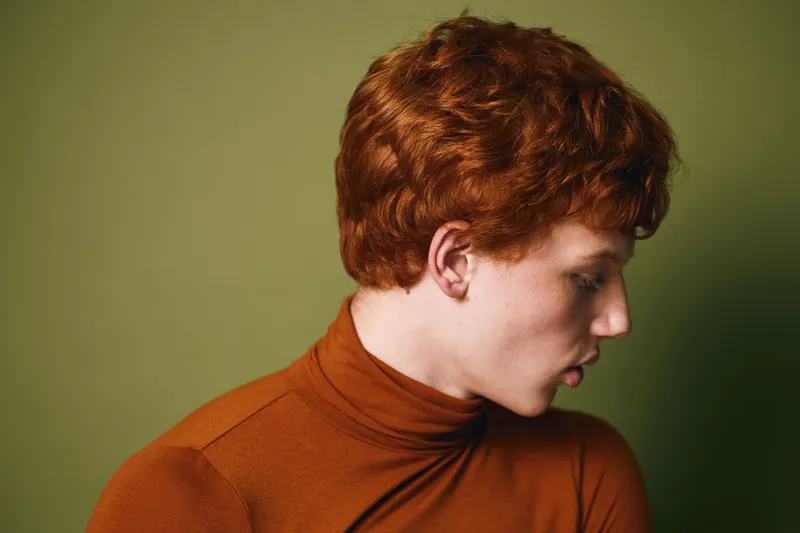 Young man with red curly hair wearing a brown turtleneck looking down against a plain olive background in studio lighting for portrait or fashion concept.
