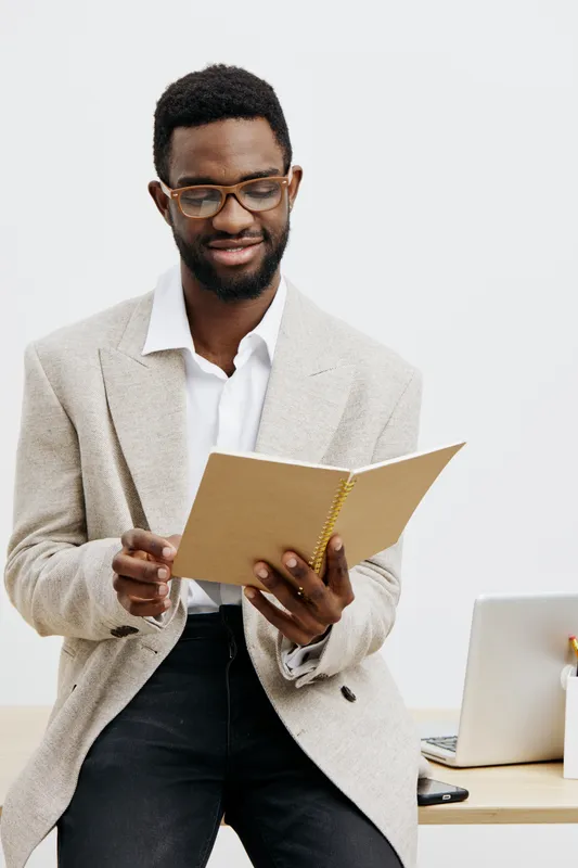 Confident and professional young African man wearing glasses and beige blazer reading a notebook while standing in front of a light background, creating a positive and focused mood, suitable for busin