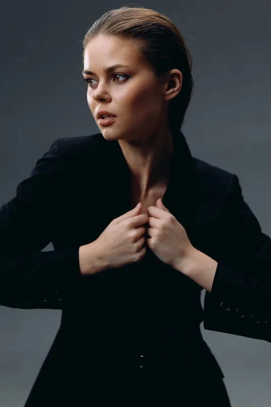 Portrait of a thoughtful woman in black suit with slicked-back hair and neutral expression in dark studio lighting.
