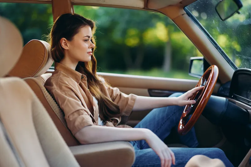 Young woman driving a vintage car, wearing casual clothes, with a serene expression in a sunny outdoor setting, conveying freedom and adventure.