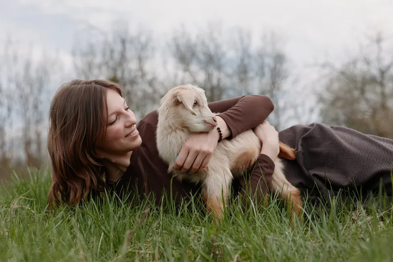 A woman lies on grass outdoors, cradling a young goat kid in her arms. Bright sky and natural light emphasize calm companionship and a simple rural lifestyle, capturing warmth, trust, and connection