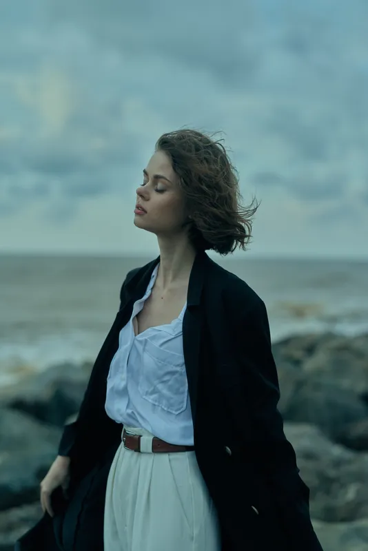 Windy beach scene with a coat over a white dress, rocky shoreline and open sea, a contemplative mood, outdoor fashion inspiration, windblown movement and elegant calm by the shore
