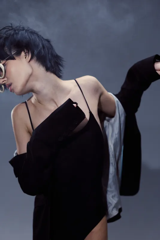 Stylish woman wearing black dress and glasses posing with dynamic movement in moody lighting.