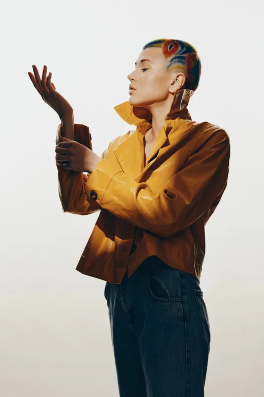 Stylish woman with colorful short hair wearing a mustard jacket and blue jeans, posing thoughtfully against a bright background in a modern fashion concept.