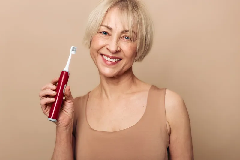 Smiling mature woman holds electric toothbrush, showing dental care routine. Beige background and casual outfit create warm, healthy lifestyle concept focused on oral hygiene and self-care.