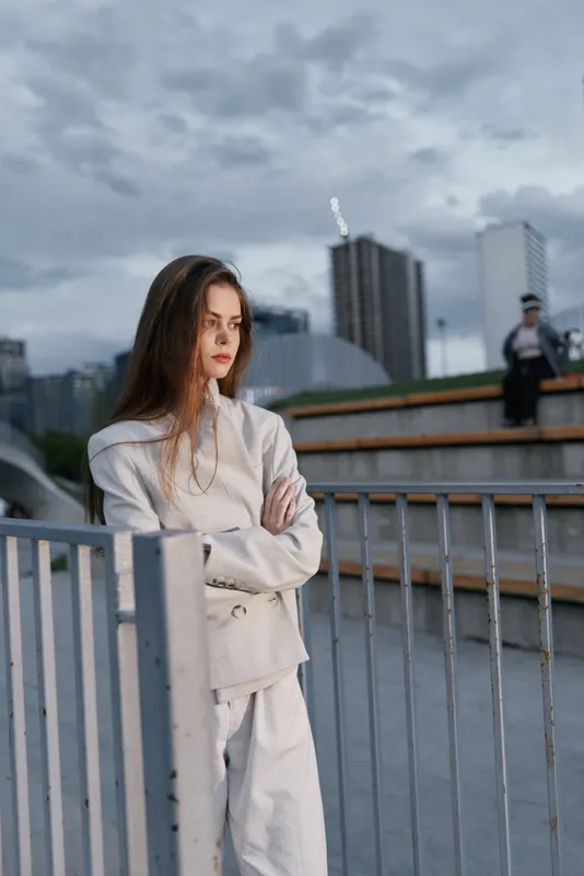 Thoughtful woman in beige outfit standing by metal fence outdoors with cityscape background cloudy sky serious mood urban fashion style