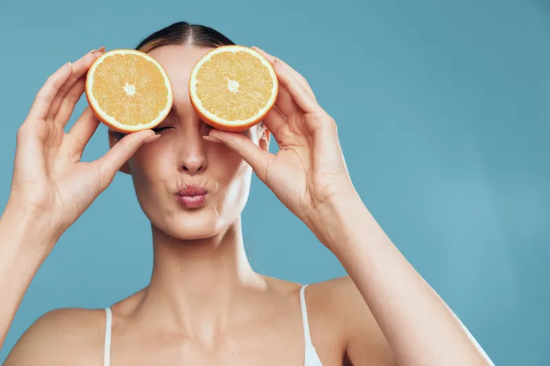 Young woman holding two halves of a grapefruit in front of her eyes, making a playful face, light blue background, casual outfit, fresh fruit concept, healthy lifestyle.