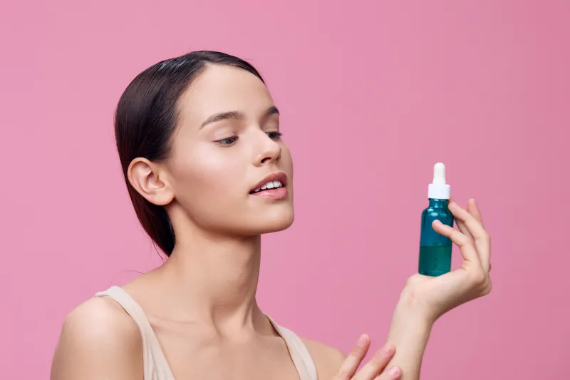 Young woman with natural makeup holding a skincare serum against a pink background, showcasing healthy skin and beauty products