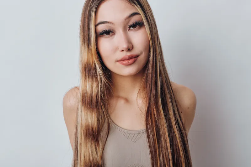 Young woman with long straight hair wearing sleeveless top posing against plain light background looking at camera with natural makeup and neutral expression indoors.