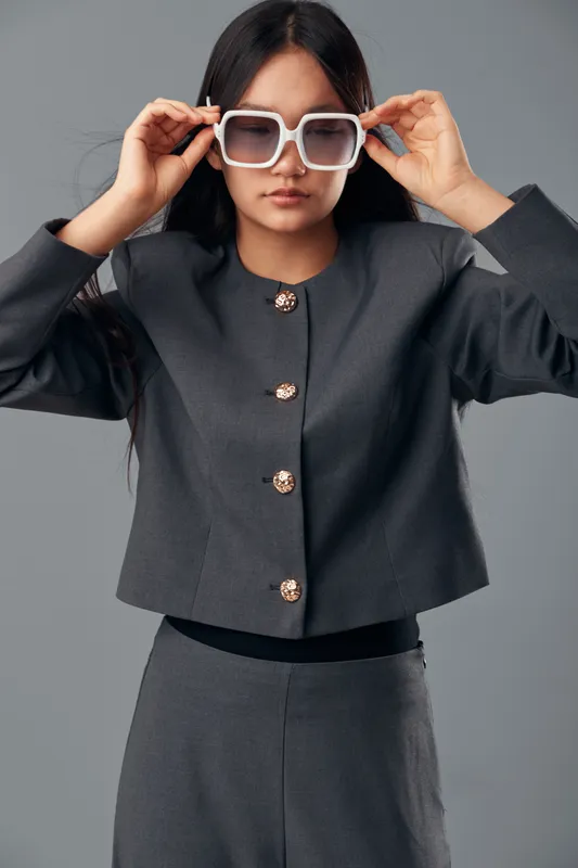 Young woman in dark gray fashionable suit adjusts oversized white sunglasses against plain background. Stylish model with long hair wears modern outfit for fashion, portrait, and lifestyle concept.
