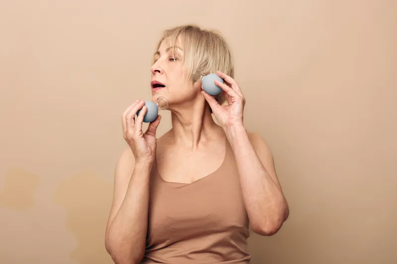 Mature woman with light hair uses facial massage rollers on skin in beige top. Close-up in neutral studio setting. Concept of anti aging, skincare routine, relaxation and beauty treatment.