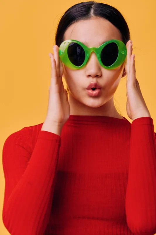 Young woman with long dark hair wearing oversized green sunglasses and red sweater poses with surprised expression on yellow background in studio portrait with fashion style.