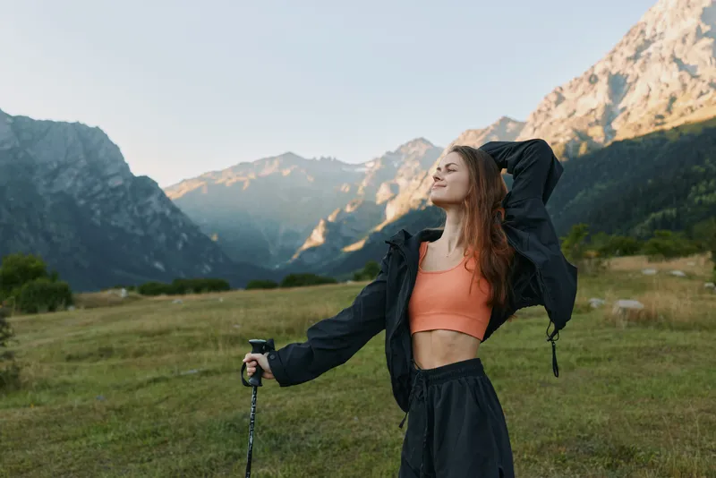 A cheerful outdoorswoman stands in a mountain meadow, wearing a coral top and dark jacket, holding trekking poles, arms raised, enjoying fresh air and bright sunshine.