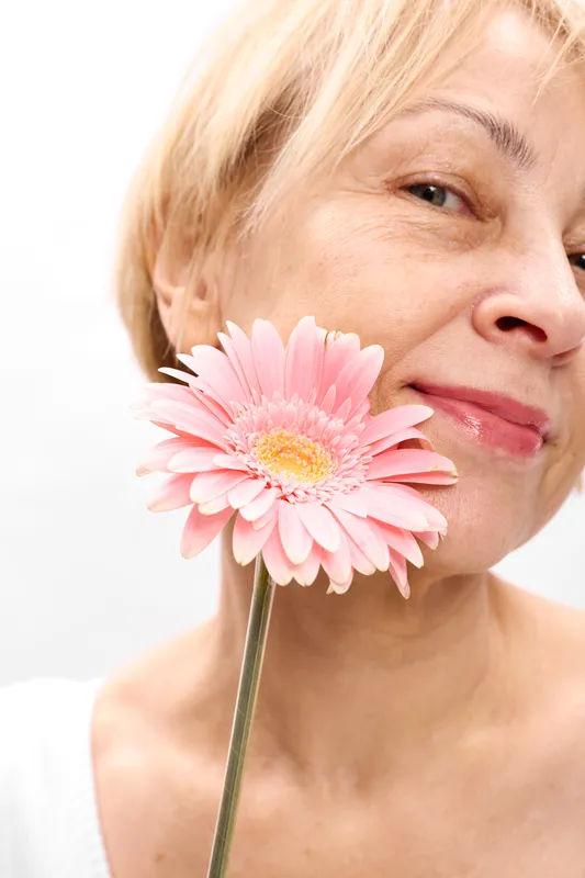 Woman portrait pink gerbera closeup smile skincare, elderly woman holding flower near cheek, soft studio light, smooth complexion, white background, peaceful expression